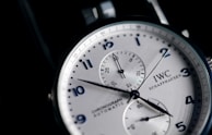A close-up view of a stylish wristwatch featuring a white dial with black numerals and two subdials. The watch is branded with 'IWC Schaffhausen' and includes the words 'Chronograph Automatic'. The hands are sleek and contrast sharply against the dial, which has a silver finish adding to the elegance of the design.