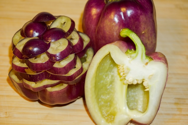 A pair of vegetables are displayed on a wooden surface, featuring a purple pepper with one half cut open to reveal its hollow interior and seeds. Another is intricately sliced to create a visually interesting pattern, adding an artistic touch.