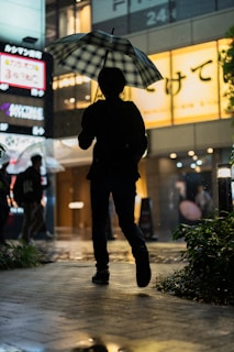 Silhouetted figure with an umbrella walking past vibrant billboards reflecting on rain-soaked streets.