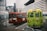 Two small buses are parked in a lot with a backdrop of modern, tall buildings. The buses have colorful exteriors, one red and one green, both featuring text in a language that uses Chinese characters. A person in a green jacket is standing nearby, with mountains visible in the distance.