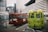 Two small buses are parked in a lot with a backdrop of modern, tall buildings. The buses have colorful exteriors, one red and one green, both featuring text in a language that uses Chinese characters. A person in a green jacket is standing nearby, with mountains visible in the distance.