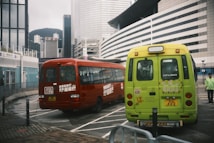 Two small buses are parked in a lot with a backdrop of modern, tall buildings. The buses have colorful exteriors, one red and one green, both featuring text in a language that uses Chinese characters. A person in a green jacket is standing nearby, with mountains visible in the distance.