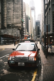 A red taxi is parked on a city street lined with tall buildings. The urban setting features a combination of older and modern architecture, with narrow streets and a tram visible in the background. Street signs, parked bicycles, and a few pedestrians are present, creating a bustling city atmosphere.