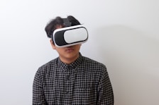 A person wearing a black and white plaid shirt uses a virtual reality headset against a plain white background.