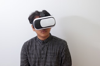 A person wearing a black and white plaid shirt uses a virtual reality headset against a plain white background.
