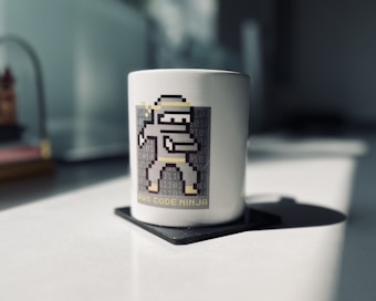 A white ceramic mug sits on a dark coaster, featuring a pixel art character resembling a ninja with a sword, accompanied by binary code in the background. The words 'AWS Code Ninja' are printed below the character. The background appears to be indoors with blurred details.