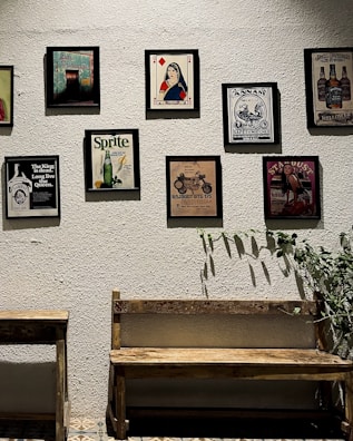 A collection of vintage-style framed posters and advertisements are arranged on a textured wall. Below the posters is a wooden bench and table, and a plant with long leaves is on the right side.
