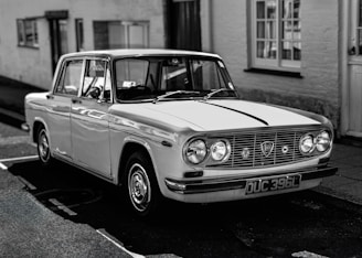 A vintage car with a classic design is parked on a street. It features a boxy shape with a chrome grille and round headlights. The car's surface reflects subtle light and shadows, evoking a sense of nostalgia. The background includes a blurred view of a building with windows, adding context to an urban setting.