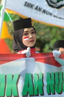 A person wearing a black hat with a gold emblem and a face painted with a red and white flag design is holding a large banner. The background features a blurred outdoor setting with a green and orange color palette.