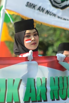 A person wearing a black hat with a gold emblem and a face painted with a red and white flag design is holding a large banner. The background features a blurred outdoor setting with a green and orange color palette.