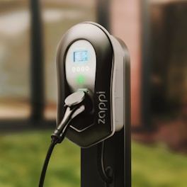 A sleek electric vehicle charging station with a black and gray design, featuring digital display and control buttons. The charger is connected, indicating it is ready for use. It is set against a blurred background, suggesting an outdoor environment.