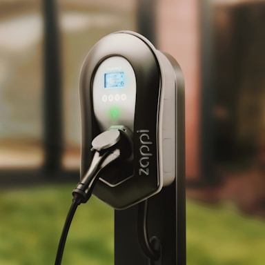 A sleek electric vehicle charging station with a black and gray design, featuring digital display and control buttons. The charger is connected, indicating it is ready for use. It is set against a blurred background, suggesting an outdoor environment.