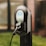 A sleek electric vehicle charging station with a black and gray design, featuring digital display and control buttons. The charger is connected, indicating it is ready for use. It is set against a blurred background, suggesting an outdoor environment.
