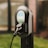 A sleek electric vehicle charging station with a black and gray design, featuring digital display and control buttons. The charger is connected, indicating it is ready for use. It is set against a blurred background, suggesting an outdoor environment.
