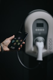 A hand holding a smartphone with a visible app interface. In the background, there is a modern electric vehicle charging station with a charger plugged in.
