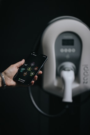 A hand holding a smartphone with a visible app interface. In the background, there is a modern electric vehicle charging station with a charger plugged in.