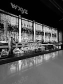 A modern bar setup features an illuminated backdrop with shelves holding various bottles. Upside-down glasses are neatly arranged along the countertop. Bright circular lights create a sparkling effect behind the bottles, contributing to a sophisticated ambiance. A tap handle with the text 'FLYING MACHINE' is visible on the left side.
