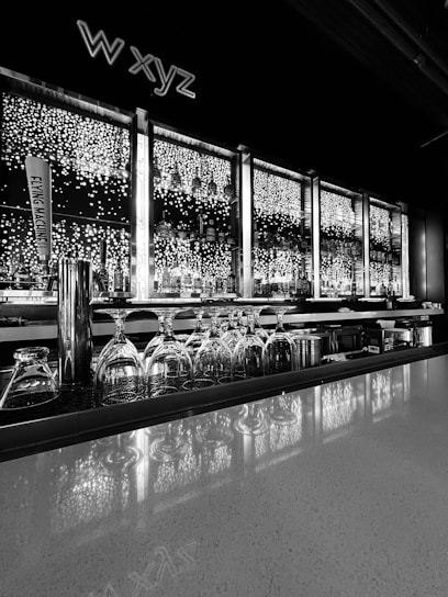 A modern bar setup features an illuminated backdrop with shelves holding various bottles. Upside-down glasses are neatly arranged along the countertop. Bright circular lights create a sparkling effect behind the bottles, contributing to a sophisticated ambiance. A tap handle with the text 'FLYING MACHINE' is visible on the left side.