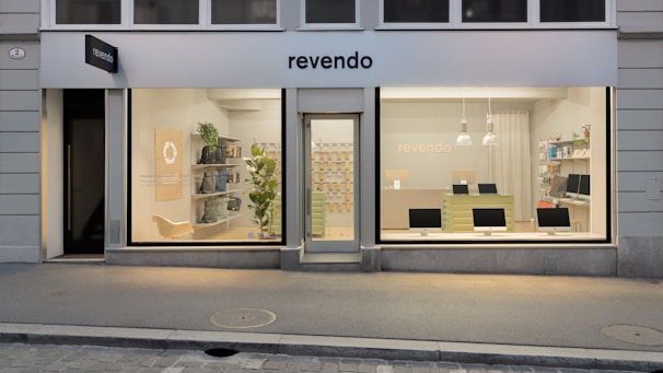 A modern storefront with large windows displaying a clean, minimalist interior. On the left, the store features a seating area with shelves holding various items and a green plant. The central door is flanked by two large glass panels showcasing multiple computer monitors on a counter. Soft lighting and a light color palette create a welcoming atmosphere.