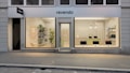 A modern storefront with large windows displaying a clean, minimalist interior. On the left, the store features a seating area with shelves holding various items and a green plant. The central door is flanked by two large glass panels showcasing multiple computer monitors on a counter. Soft lighting and a light color palette create a welcoming atmosphere.