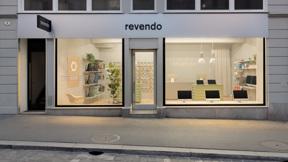 A modern storefront with large windows displaying a clean, minimalist interior. On the left, the store features a seating area with shelves holding various items and a green plant. The central door is flanked by two large glass panels showcasing multiple computer monitors on a counter. Soft lighting and a light color palette create a welcoming atmosphere.