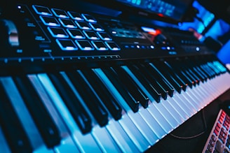 A close-up view of an electronic keyboard featuring black and white keys, illuminated by blue lighting. Additional control pads and knobs are visible on the upper part, suggesting a music production device. The scene has a futuristic and technological atmosphere.