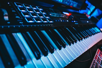 A close-up view of an electronic keyboard featuring black and white keys, illuminated by blue lighting. Additional control pads and knobs are visible on the upper part, suggesting a music production device. The scene has a futuristic and technological atmosphere.