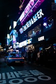 Neon signs illuminate a bustling street at night, with a lively atmosphere as people walk along the sidewalk. The vibrant signage includes words like 'CHICKEN & BEER' and 'LUSH,' creating a colorful scene. A sleek car is parked on the street, adding a modern touch.