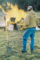 A person wearing a brown jacket and blue jeans stands on a grassy field, observing a large fire consisting of wooden debris and household items. A shovel is positioned upright in the ground next to the person, and the background features leafless trees.
