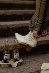 A pair of white cowboy boots is worn by someone sitting on stone steps. Two bottles of Corona Extra beer are placed on the step, supported by a cardboard holder. The person is wearing a long-sleeved greenish-brown top and adjusting their boots.