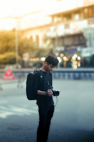 A young person stands in a street setting, holding a camera and looking down, wearing a black backpack and casual black clothing. The background is blurred, showing hints of urban elements with some bokeh lights and indistinct buildings.