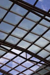 A glass ceiling with a crisscross pattern of metal supports is visible against a partly cloudy sky. The structure allows natural light to filter through, creating an open and airy atmosphere.