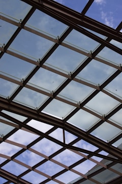 A glass ceiling with a crisscross pattern of metal supports is visible against a partly cloudy sky. The structure allows natural light to filter through, creating an open and airy atmosphere.