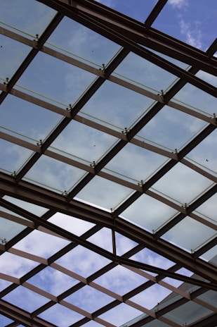 A glass ceiling with a crisscross pattern of metal supports is visible against a partly cloudy sky. The structure allows natural light to filter through, creating an open and airy atmosphere.