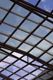 A glass ceiling with a crisscross pattern of metal supports is visible against a partly cloudy sky. The structure allows natural light to filter through, creating an open and airy atmosphere.