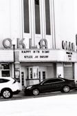 A vintage cinema building with marquee signage displays a birthday message for Kylee Jo Busby. The facade features large vertical windows and art deco style lettering spelling 'OKLA'. Parked cars are visible in front of the building.