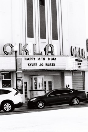 A vintage cinema building with marquee signage displays a birthday message for Kylee Jo Busby. The facade features large vertical windows and art deco style lettering spelling 'OKLA'. Parked cars are visible in front of the building.
