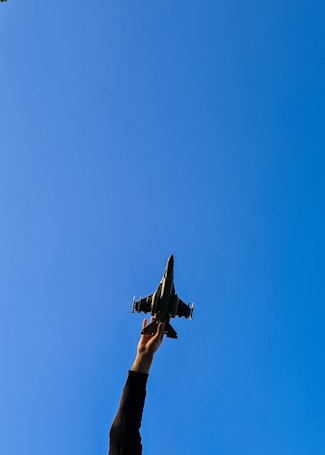 A person extends their arm upwards, holding a model aircraft against a clear blue sky. The silhouette of the aircraft and the person’s hand are in sharp contrast with the bright sky.