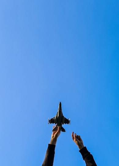 Hands reaching upward hold a model fighter jet against a clear blue sky, conveying a sense of aspiration or imagination.
