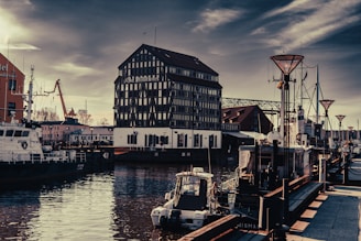 A waterfront scene with an older-style building prominently displaying the name on it. The setting features several docked boats and ships in calm waters, surrounded by both traditional and industrial architecture. There are street lamps and a pedestrian walkway alongside the water, and an overcast sky in the background.