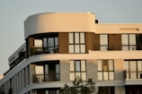 A modern apartment building with large windows and balconies, featuring a mix of curved and straight architectural lines with multiple levels. The facade includes contrasting materials and colors, likely brick and concrete, and there are some potted plants visible on the balconies.