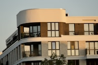 A modern apartment building with large windows and balconies, featuring a mix of curved and straight architectural lines with multiple levels. The facade includes contrasting materials and colors, likely brick and concrete, and there are some potted plants visible on the balconies.