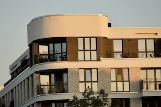 A modern apartment building with large windows and balconies, featuring a mix of curved and straight architectural lines with multiple levels. The facade includes contrasting materials and colors, likely brick and concrete, and there are some potted plants visible on the balconies.