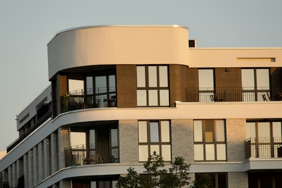 A modern apartment building with large windows and balconies, featuring a mix of curved and straight architectural lines with multiple levels. The facade includes contrasting materials and colors, likely brick and concrete, and there are some potted plants visible on the balconies.