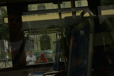 Reflections are seen through a bus window, showing two people outside. One person is wearing a white shirt and the other has a red headscarf. The background includes a building with large windows and architectural details.