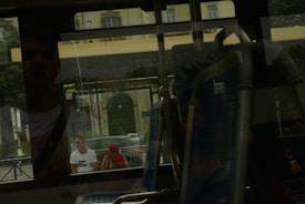 Reflections are seen through a bus window, showing two people outside. One person is wearing a white shirt and the other has a red headscarf. The background includes a building with large windows and architectural details.