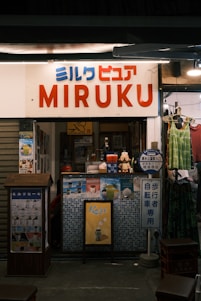 A small shop with a sign saying 'MIRUKU' in large letters and Japanese text above. The shop has various advertisements and posters on the front, showcasing drinks and desserts. A mannequin or figure is visible near the entrance, and a couple of hanging clothes are on display outside. The interior is dimly lit, adding a cozy atmosphere.