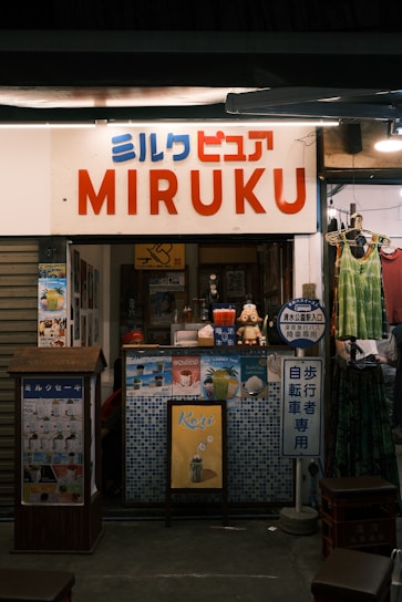 A small shop with a sign saying 'MIRUKU' in large letters and Japanese text above. The shop has various advertisements and posters on the front, showcasing drinks and desserts. A mannequin or figure is visible near the entrance, and a couple of hanging clothes are on display outside. The interior is dimly lit, adding a cozy atmosphere.