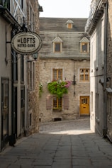 A charming narrow alleyway lined with historic stone buildings. The path leads to a picturesque house with a sloped gray roof, small dormer windows, and a bright yellow door. Flowering plants cascade from window boxes adding a touch of color. A sign for 'Les LOFTS NOTRE-DAME' hangs on one of the buildings.