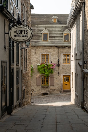 A charming narrow alleyway lined with historic stone buildings. The path leads to a picturesque house with a sloped gray roof, small dormer windows, and a bright yellow door. Flowering plants cascade from window boxes adding a touch of color. A sign for 'Les LOFTS NOTRE-DAME' hangs on one of the buildings.